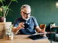 A mature man sitting in a cafe, reading on a digital tablet and drinking a cup of coffee.