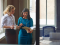 two businesswomen standing and looking at paperwork in an office with a coffee cup in hand and laptop on the table