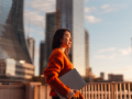 Businesswoman carrying laptop, looking at view in financial district at sunset.