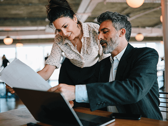 seated business man pointing to information on paper for standing female colleague