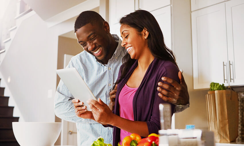 standing couple in home kitchen looking at investments on tablet