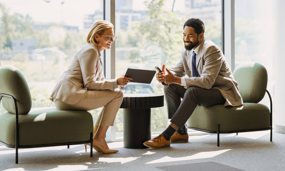 businesswoman with tablet talking with a young man, both seated on modern chairs in front of window