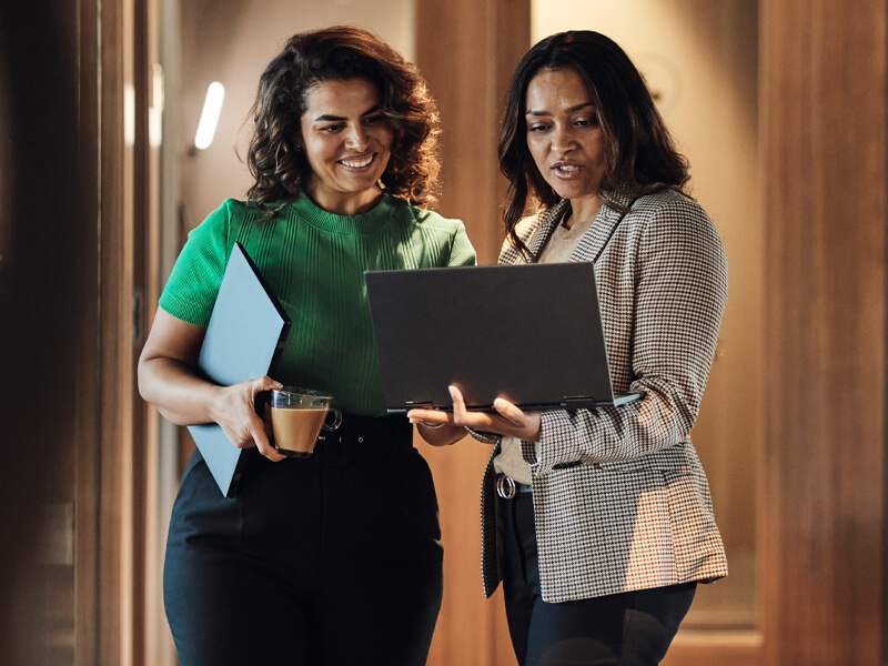 two smiling business women standing, each holding a laptop, one open