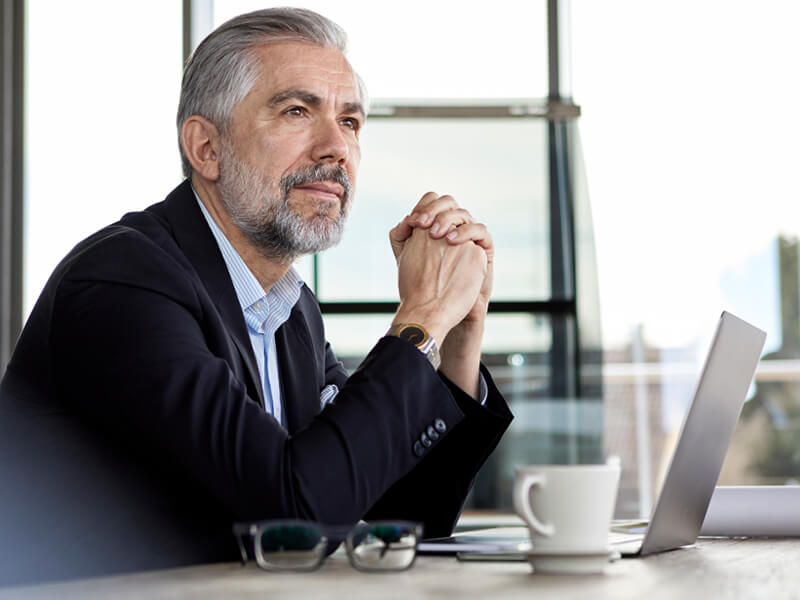 older businessman, elbows on desk, hands clasped, sitting in front of laptop