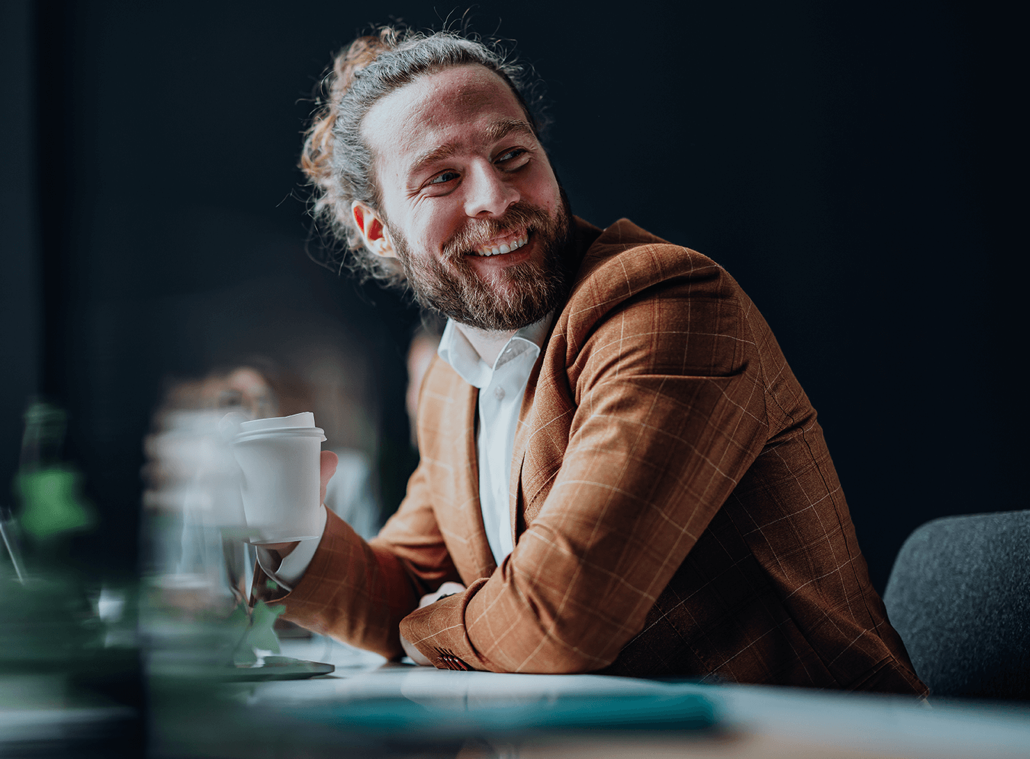 seated bearded man in rust suit jacket with coffee cup in hand