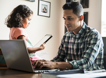  seated father working from home on laptop with daughter with tablet