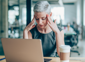 grey-haired caucasian woman with fingers to temples in front of laptop