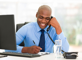 African American man sitting at desk in front of computer, writing and on phone image