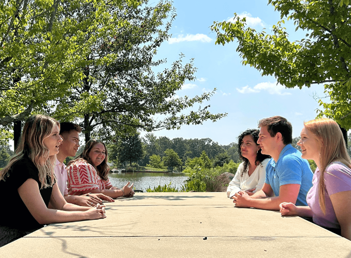 young adults sitting outside at picnic table in front of lake