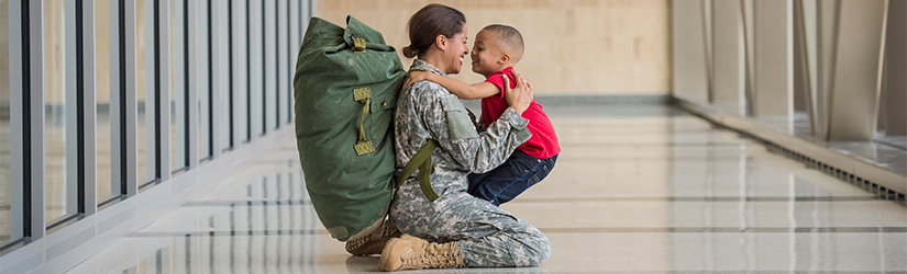 military woman in fatigues kneeling in industrial hallway hugging child